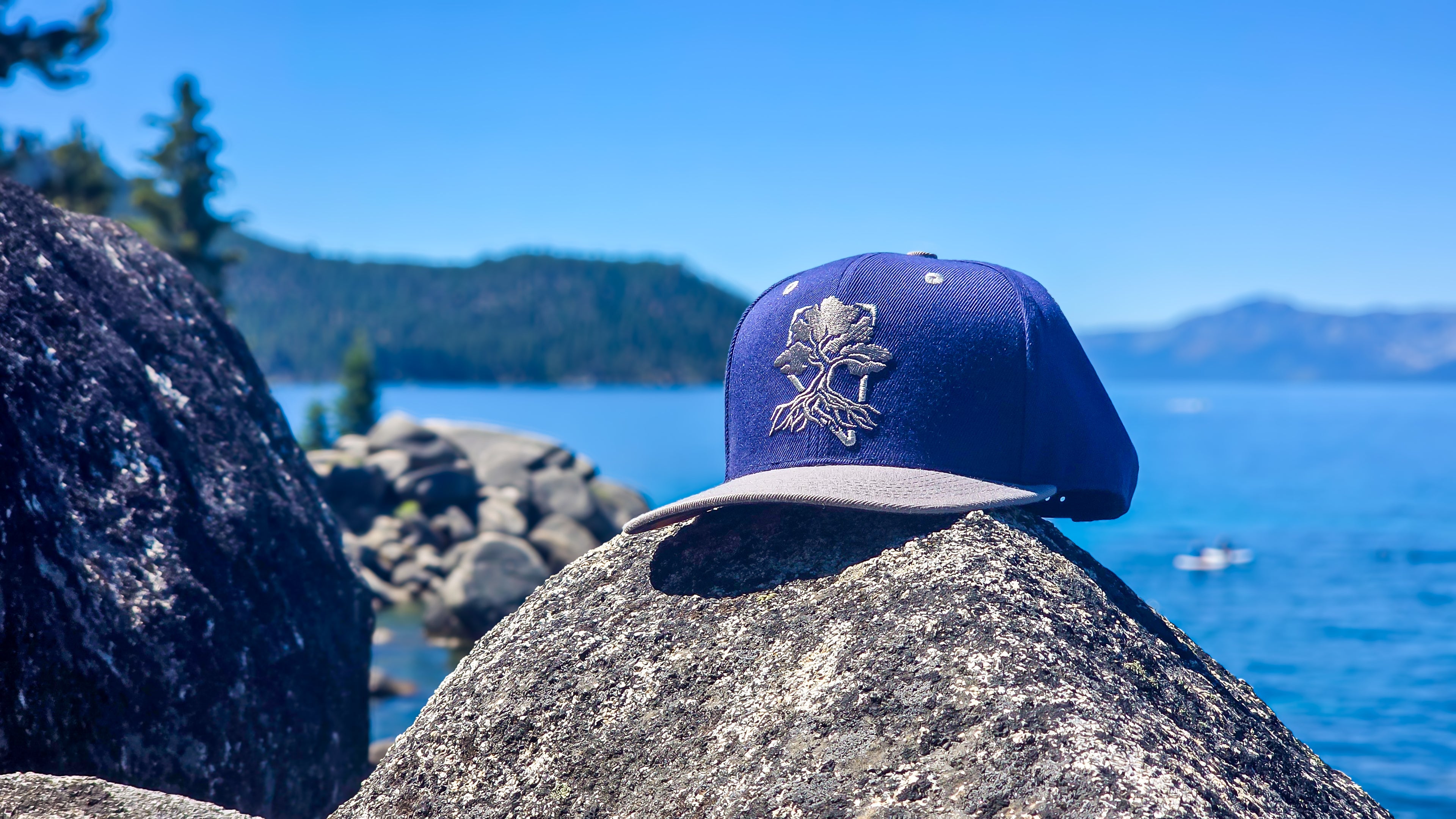 Rooted Sunset Snapback limited edition snapback hat with embroidered tree logo displayed on a rock beside Lake Tahoe